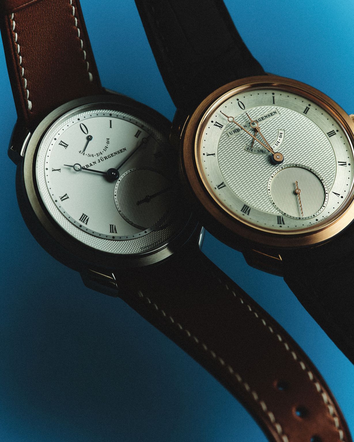 Two Urban Jürgensen wristwatches with leather straps, featuring classic Roman numeral dials and guilloché detailing, displayed against a blue background.
