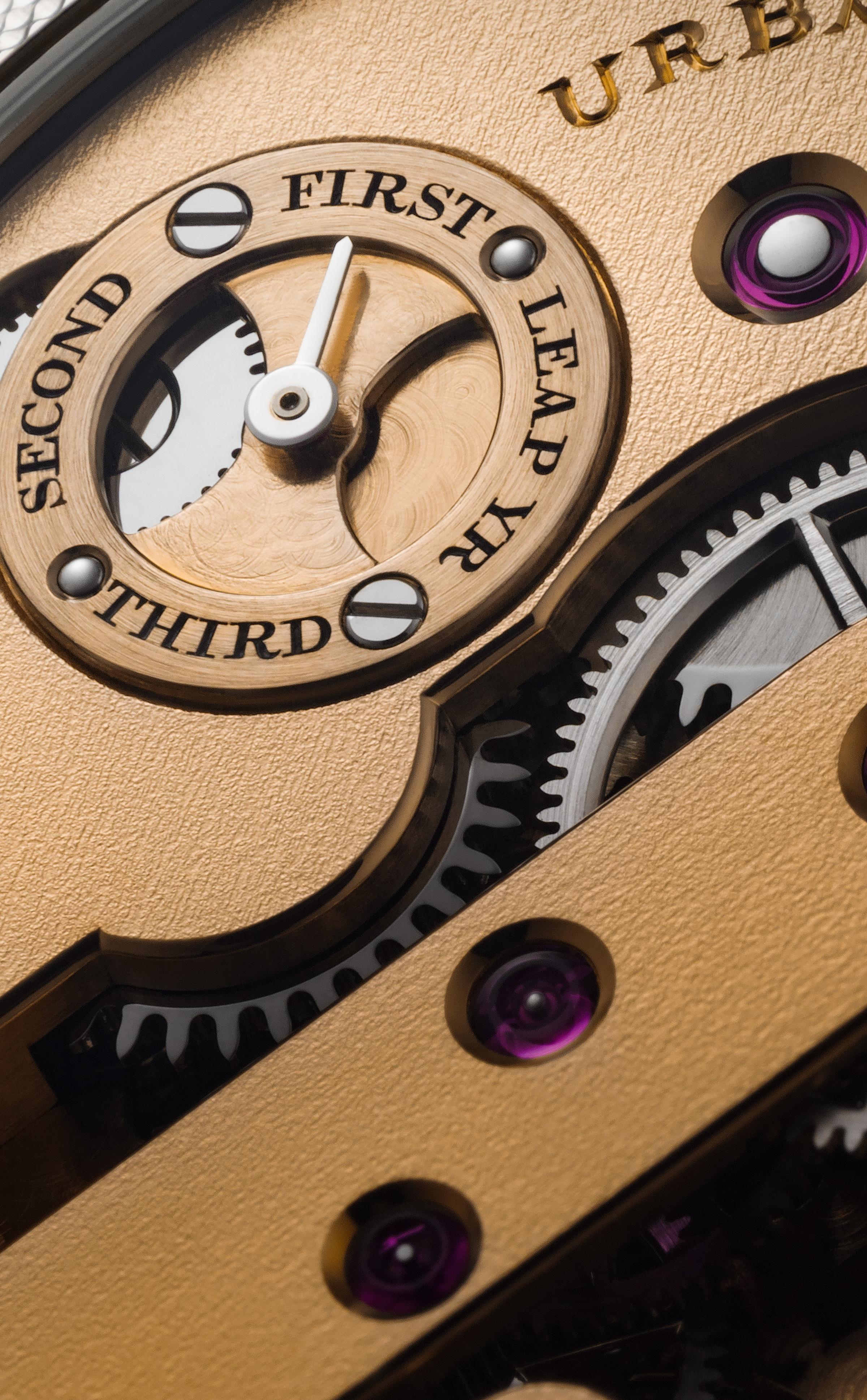 Close-up of a rose gold leap year indicator on a watch movement, showing engraved labels for First, Second, Third, and Leap Yr, with visible gears and jewel bearing.