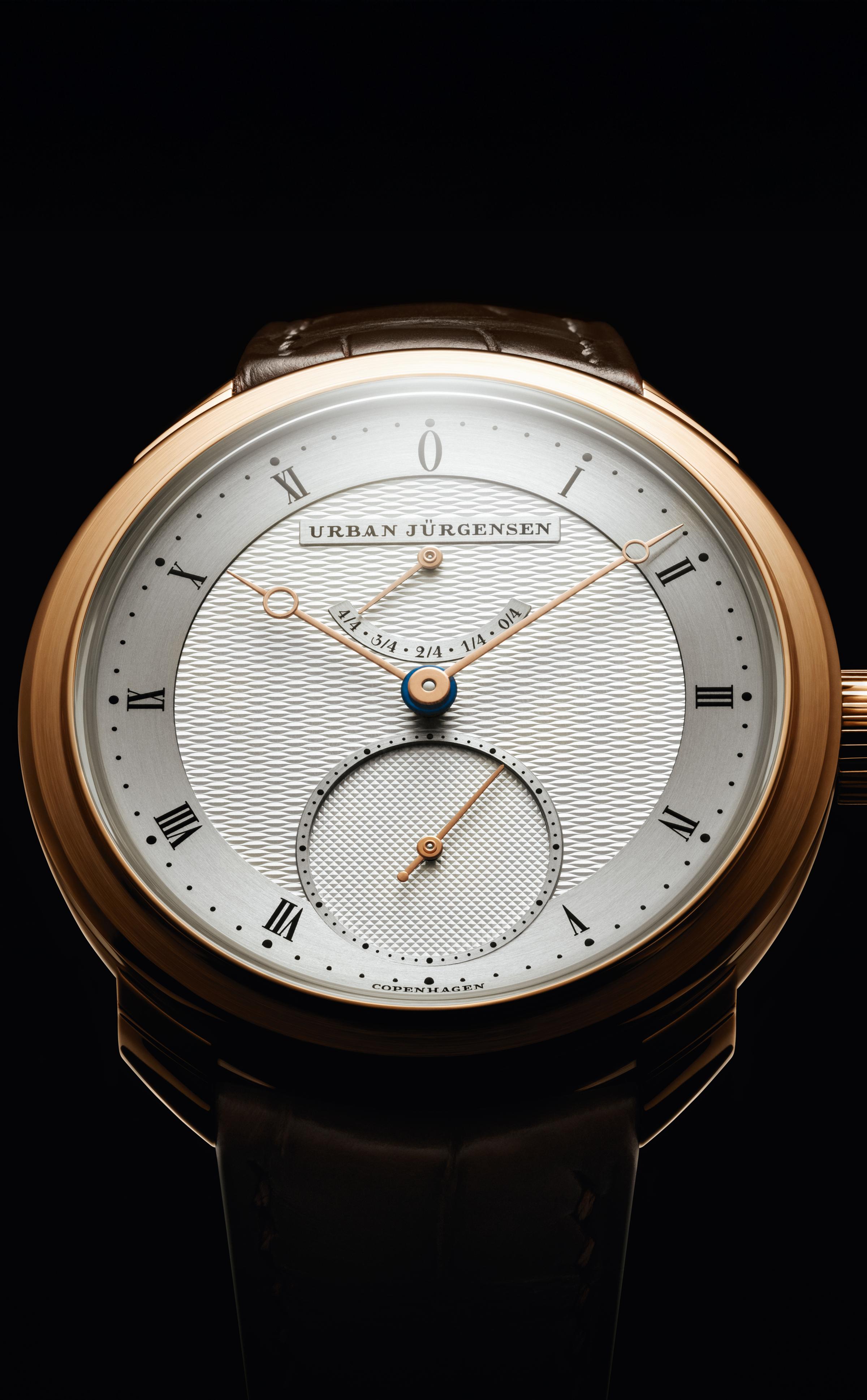 Front view of a rose gold Urban Jürgensen watch with silver guilloché dial, Roman numerals, and small seconds subdial, set against a black background.