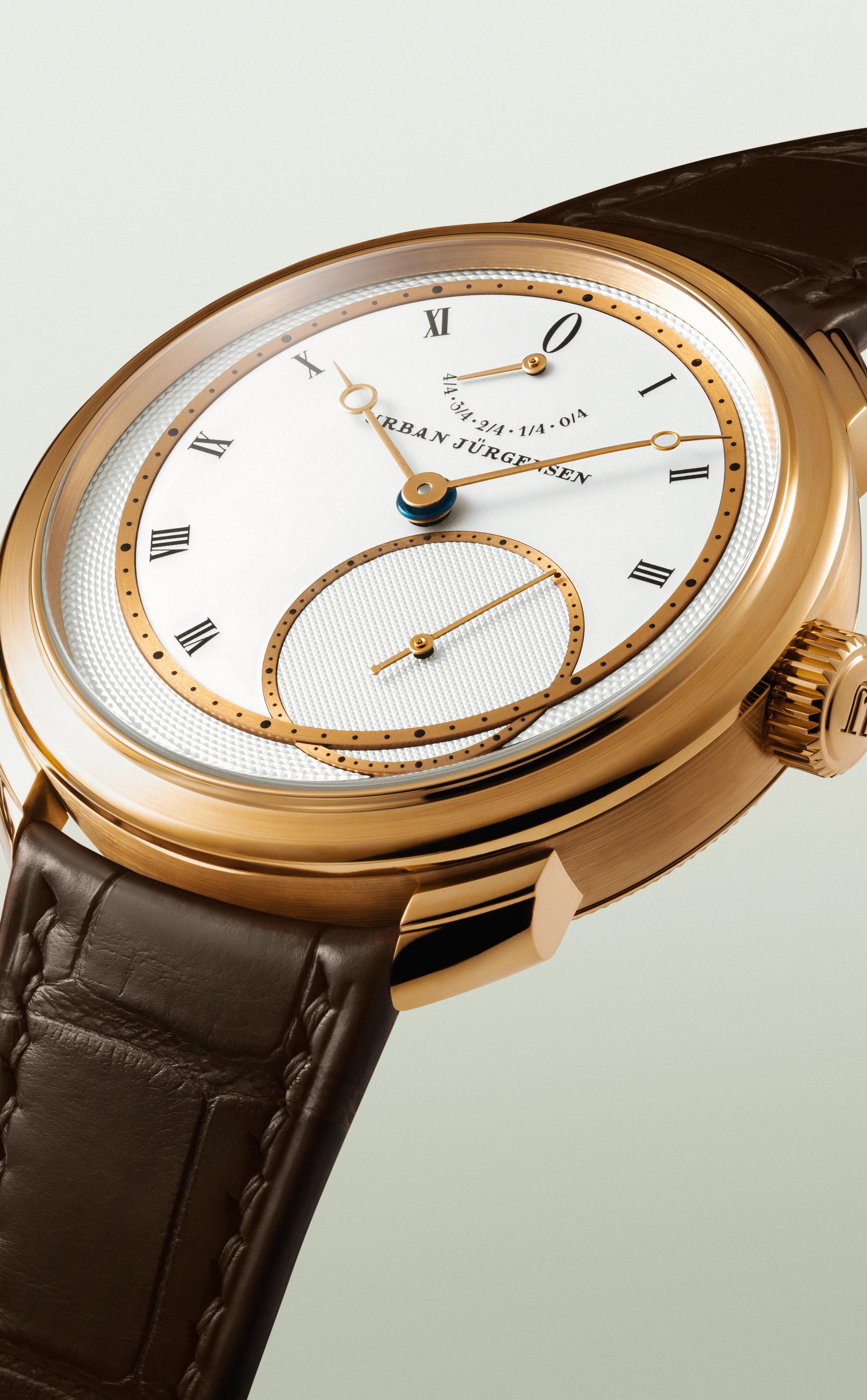 Rose gold Urban Jürgensen wristwatch with silver dial, gold hands, Roman numerals, power reserve indicator, and brown leather strap.