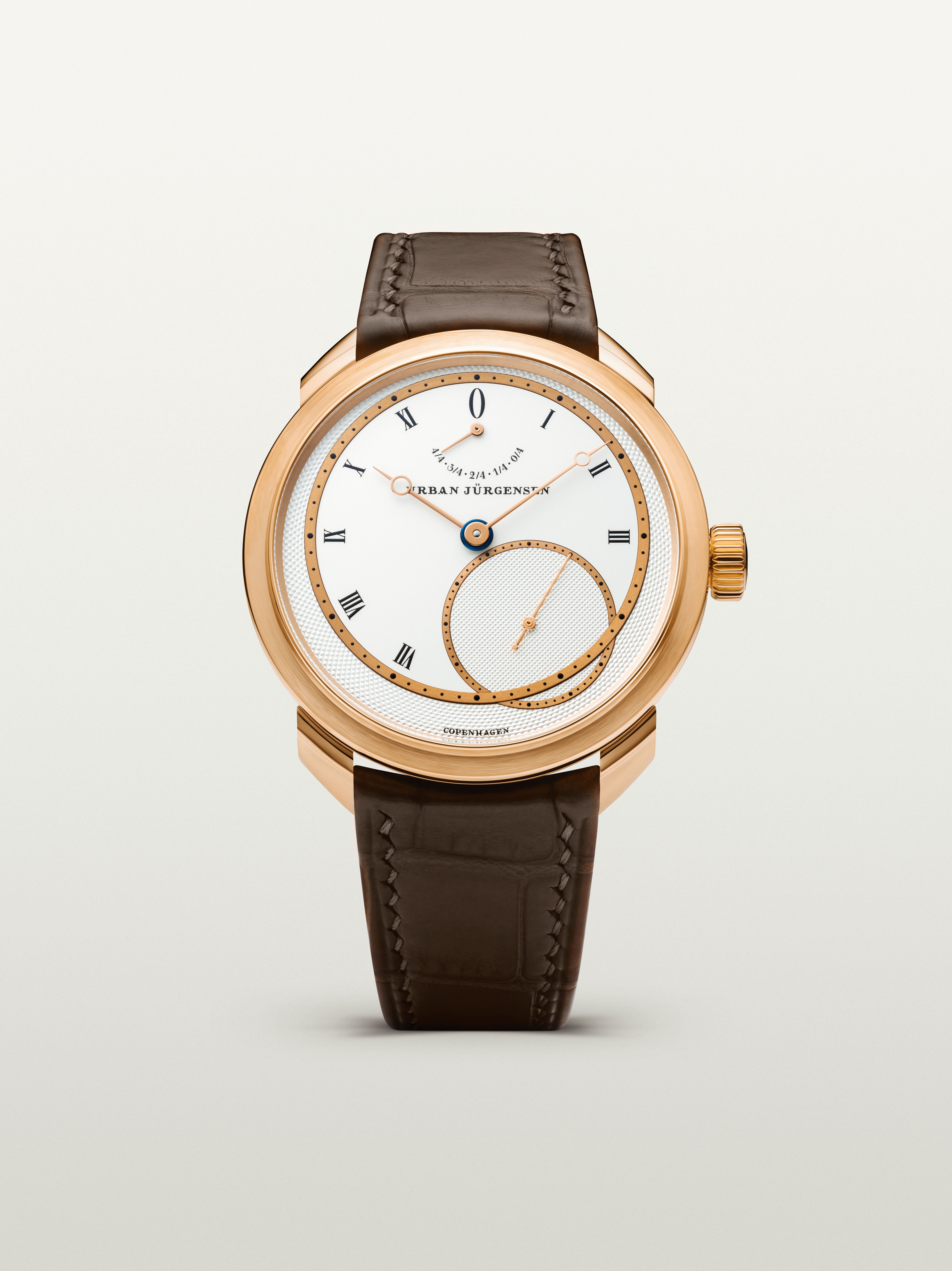 Rose gold Urban Jürgensen wristwatch with silver dial, gold hands, Roman numerals, power reserve indicator, and brown leather strap.