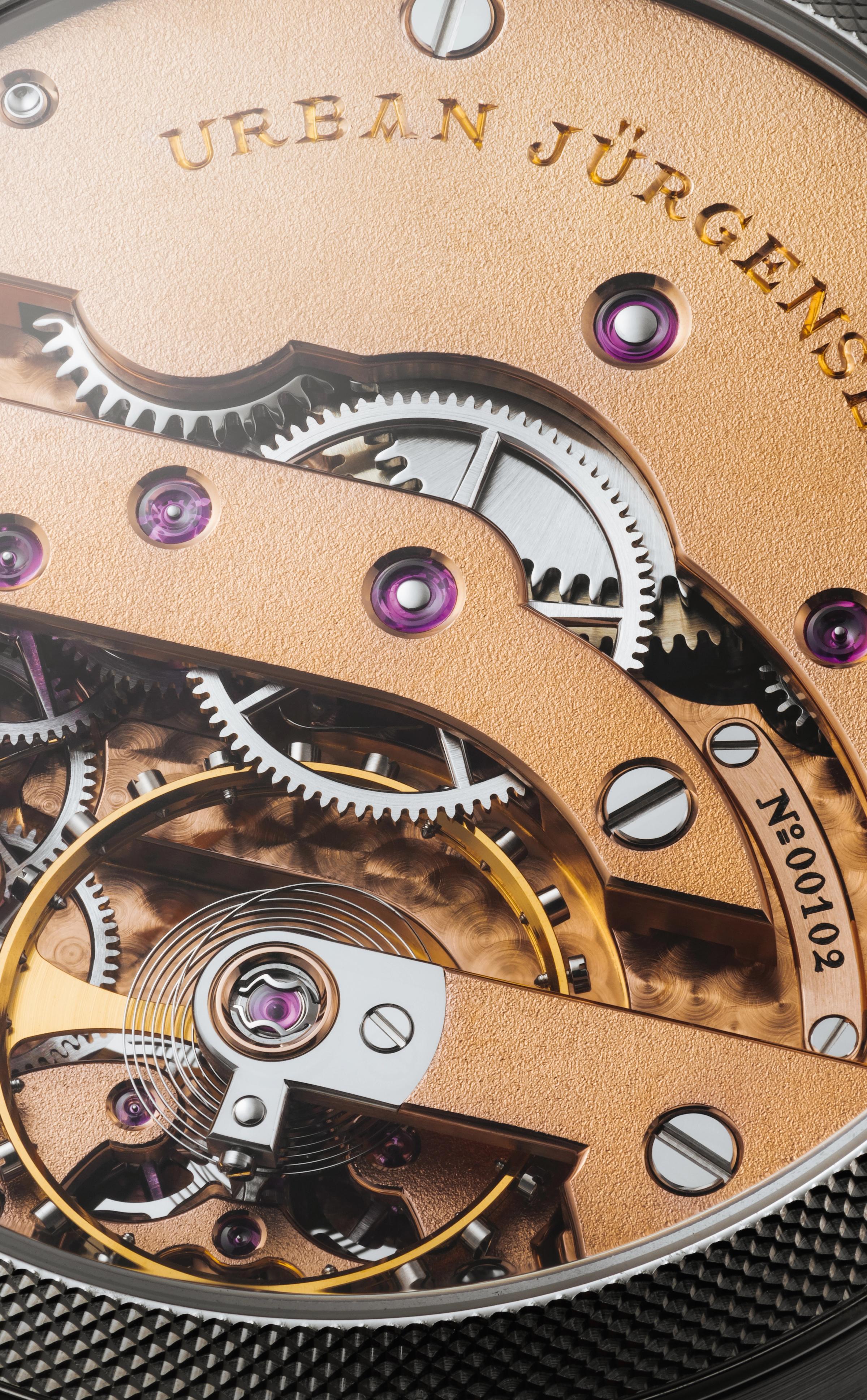 Close-up of a finely finished Urban Jürgensen watch movement showing gold-toned bridges, polished gears, ruby jewel bearings, and engraved serial number.
