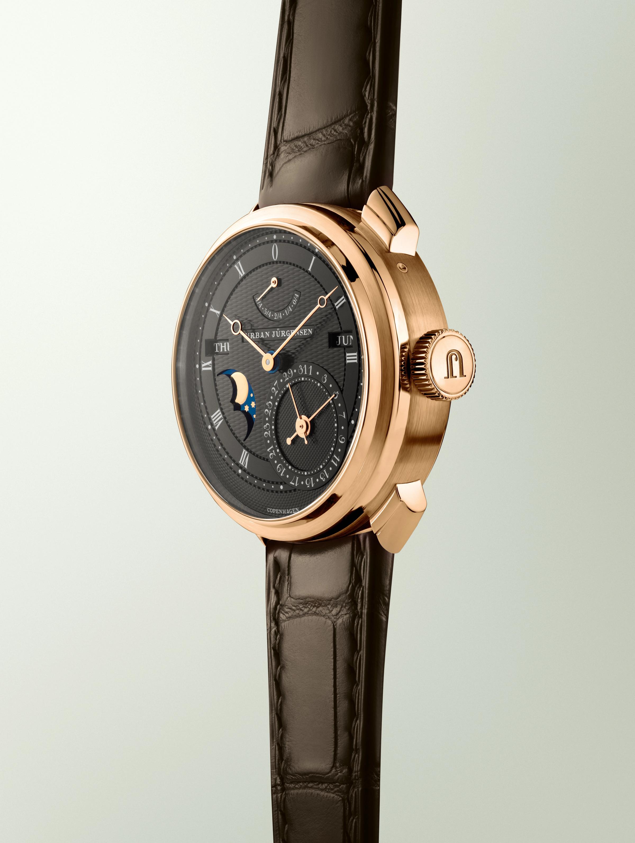 Rose gold Urban Jürgensen wristwatch with dark guilloché dial, moonphase, day and date indicators, power reserve display, and brown leather strap.
