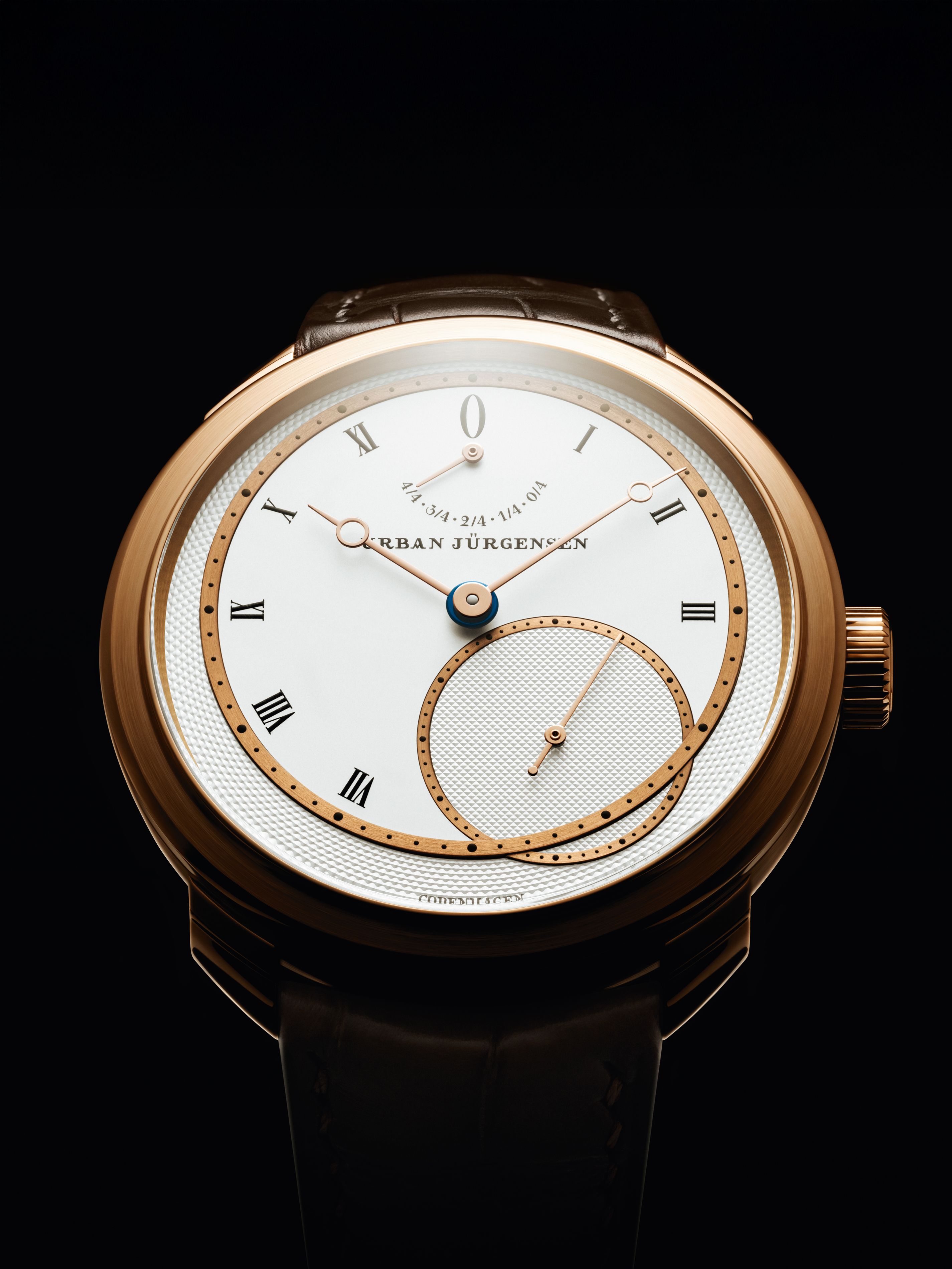Front view of a rose gold Urban Jürgensen wristwatch with silver dial, gold hands, Roman numerals, power reserve indicator, and small seconds subdial, set against a black background.
