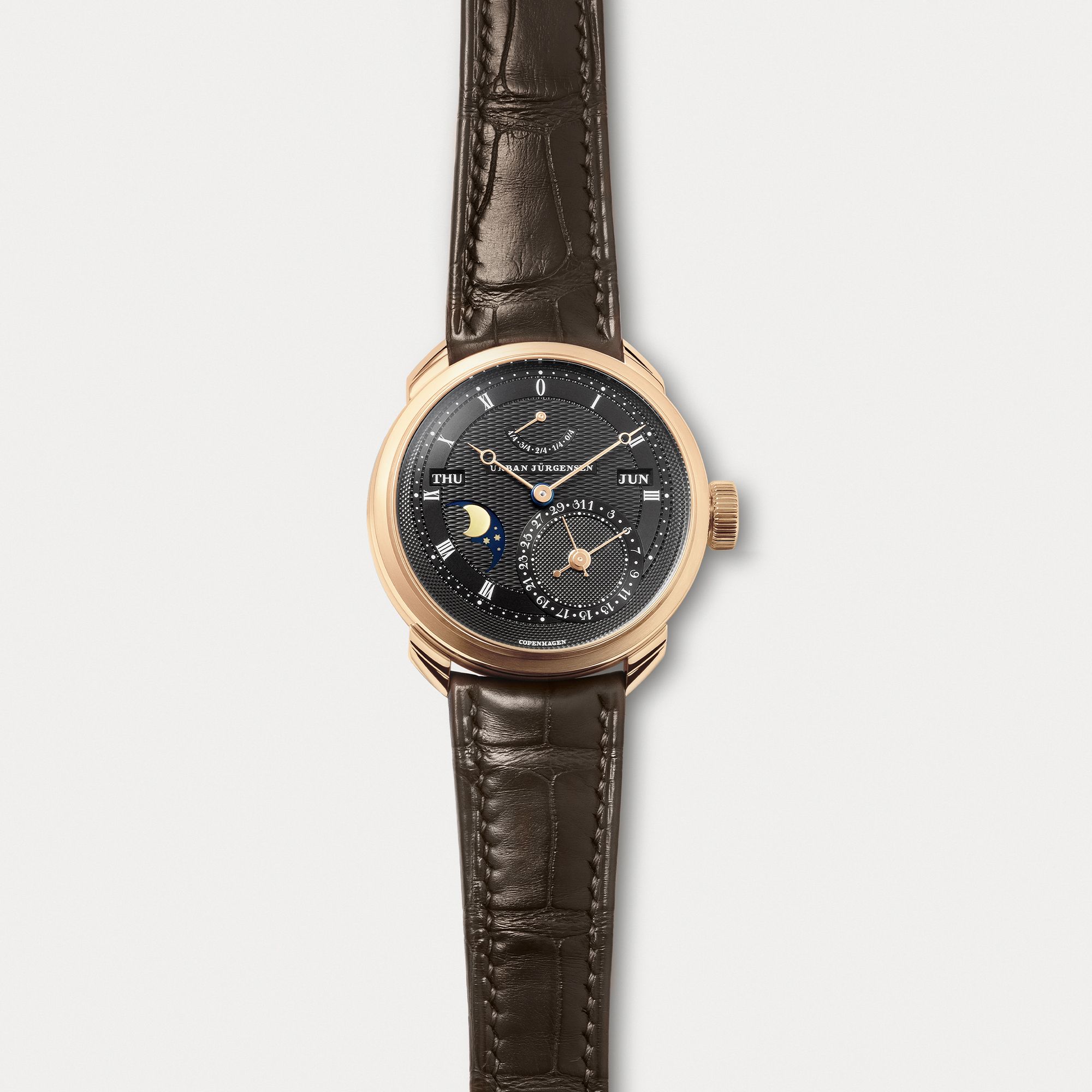 Rose gold Urban Jürgensen wristwatch with dark guilloché dial, moonphase, day and date indicators, power reserve display, and brown leather strap.