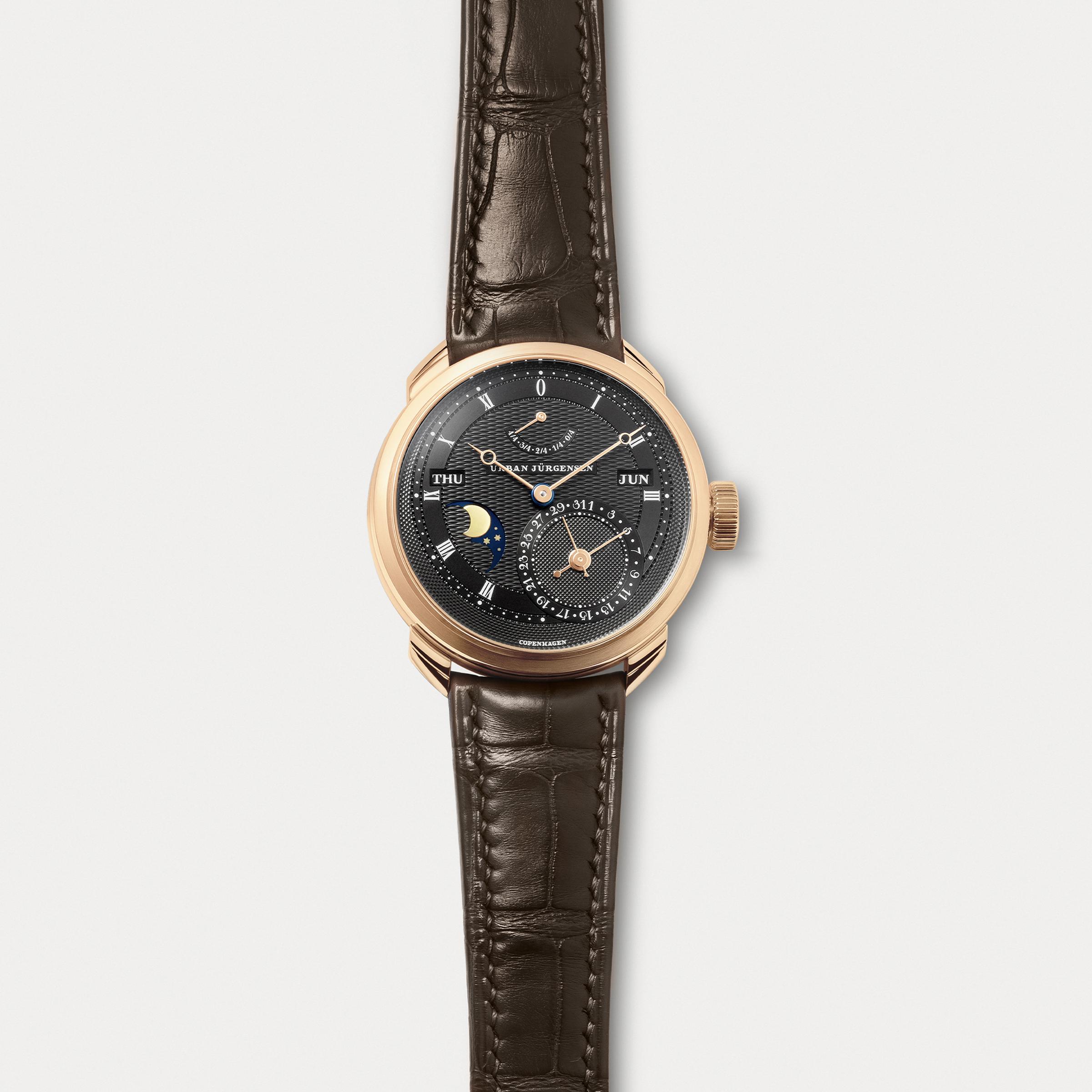Rose gold Urban Jürgensen wristwatch with dark guilloché dial, moonphase, day and date indicators, power reserve display, and brown leather strap.