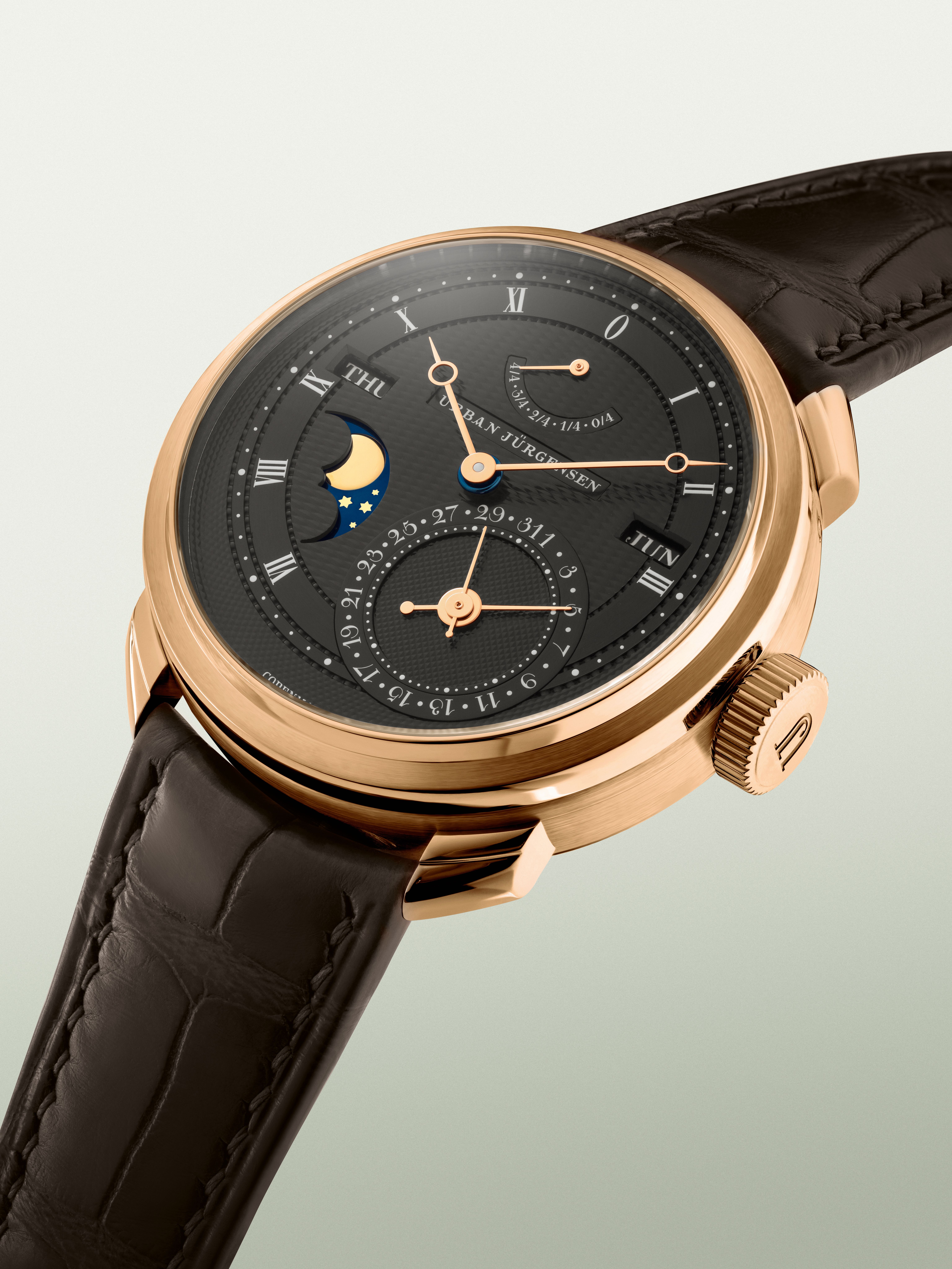 Rose gold Urban Jürgensen wristwatch with dark guilloché dial, moonphase, day and date indicators, power reserve display, and brown leather strap.