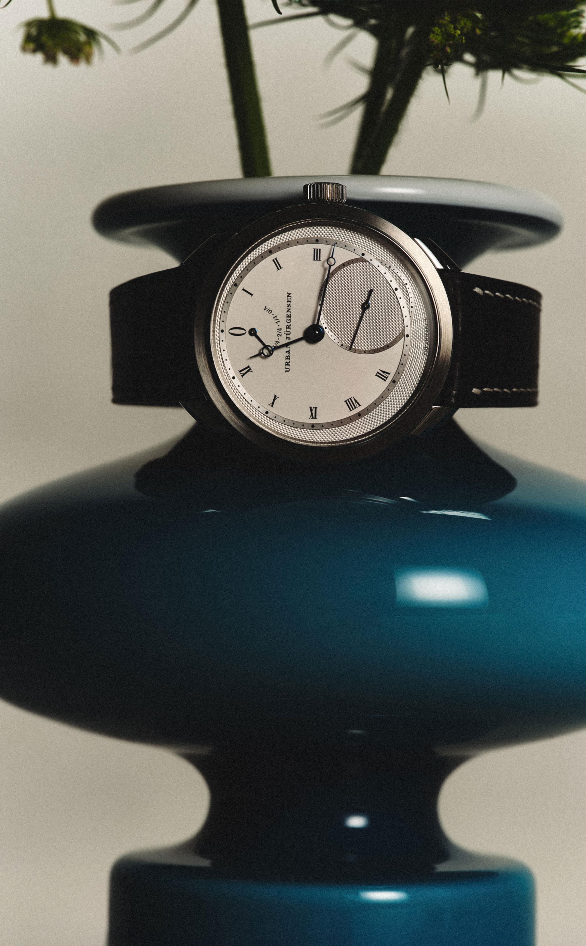 Urban Jürgensen wristwatch with silver dial and black strap displayed on a glossy dark teal vase, with green flower stems emerging from the top.